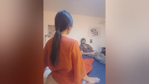 A video shows a woman in a red robe and ponytail sitting on a bed, facing a man in a checkered shirt, who is sitting cross-legged on a blue bedspread. The room has beige walls with abstract art.