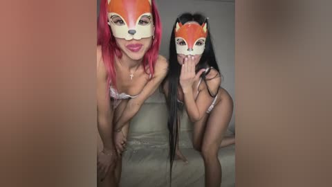 Video of two women with long hair, one with red and the other with black, wearing orange fox masks, lingerie, and posing provocatively on a couch.