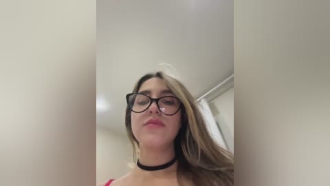 Media: A video of a young woman with fair skin, long blonde hair, and glasses, wearing a black choker necklace and a red lace top, captured from a low angle in a room with beige walls.