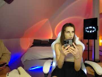 Media: Video of a young woman with light skin and brown hair, wearing a white top and black skirt, seated in a gaming chair, holding a smartphone, in a dimly lit room with orange and blue lighting and a Batman lamp.