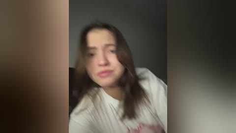 A blurry video of a young woman with straight, dark hair, wearing a white shirt, taken from a close-up angle with a dark background.