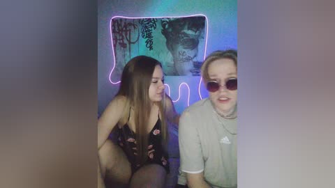 Video of two young women in a dimly lit room. One wears a floral-patterned black lingerie, the other a beige shirt. Background includes a graffiti-covered wall and a neon light strip.