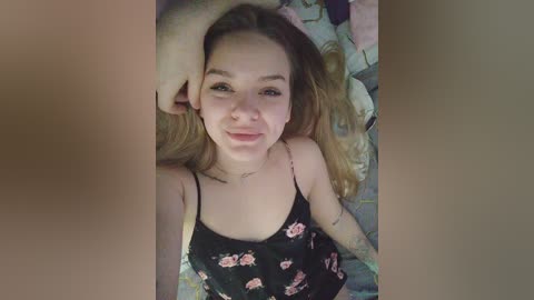 Video of a young Caucasian woman with light skin, long blonde hair, wearing a black floral tank top, lying on a bed with a colorful quilt. She has a slight smile and is looking at the camera.