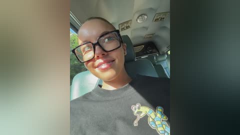 Media: Video of a smiling young woman with light skin, wearing glasses, black T-shirt with a green frog graphic, and seated in a car with beige interior, sunlight streaming in.