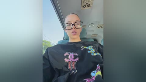 Media: Video of a young woman with fair skin, glasses, and dark hair, wearing a black shirt with colorful cartoon characters, sitting in a car with a blurred view of trees outside.