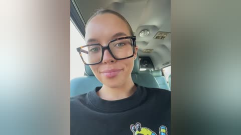 Media: A video of a smiling young woman with light skin and brown hair, wearing black-framed glasses and a black T-shirt with a colorful cartoon character, seated in a blue upholstered car seat.