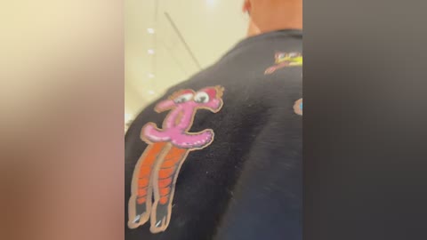 Media: Video of a person's back in a dark room, wearing a black t-shirt with a cartoonish, pink octopus graphic and red tentacles, partially blurred background.