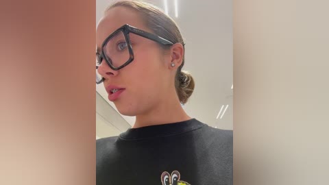 Media: Video of a fair-skinned woman with blonde hair pulled back, wearing black-framed glasses and a black shirt with a colorful logo. The background is a plain, light-colored indoor setting.