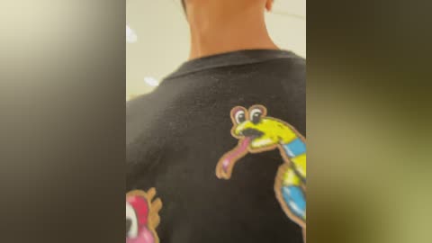 Media: Video of a person's back, wearing a black T-shirt with a colorful, cartoonish snake design. The background is blurred, with a gradient of green and yellow hues.