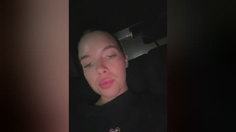 Media: A dimly lit video of a young woman with light skin and straight hair, resting her head against a black car headrest. She wears a black top.