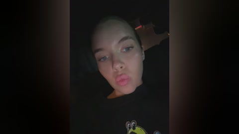 Media: A dimly lit video of a young woman with fair skin and a bald head, wearing a black shirt with a yellow cartoon character, captured from a low angle.