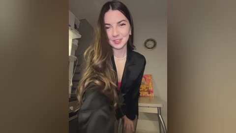 Media: Video of a smiling woman with long, wavy brown hair, wearing a black blazer, standing in a dimly lit kitchen with beige walls and a clock.