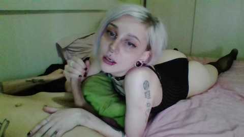 Media: Video of a young woman with platinum blonde hair, light skin, and tattoos, lying on a bed in black lingerie, holding a knife.