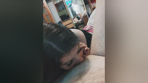 Video of a woman with short, dark hair, glasses, and a black top, lying face down on a bed, partially obscured by a person's legs. The room features wooden furniture and a TV in the background.