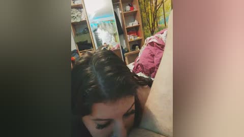Video of a young woman with dark hair and fair skin, wearing a pink shirt, leaning on a beige wall in a dimly lit room with wooden shelves, green plants, and a blue tent.