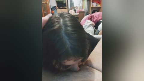 Video of a man with short dark hair performing oral sex on a man's penis. Background shows a messy room with a bed, wooden shelves, and a mirror.