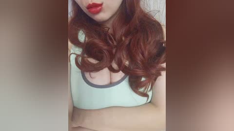 Video of a fair-skinned woman with long, wavy auburn hair, wearing a low-cut, white tank top that reveals ample cleavage, with bold red lipstick.