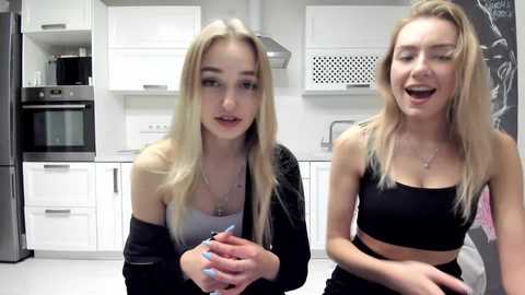 Media: Video of two young women with long blonde hair, wearing black tank tops and jackets, smiling in a modern kitchen with white cabinets, stainless steel appliances, and a blackboard wall.