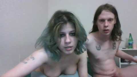 Media: Video of a naked young woman with shoulder-length, green hair and tattoos, and a young man with long brown hair, sitting on a bed in a simple room with a green bottle and a white wall.