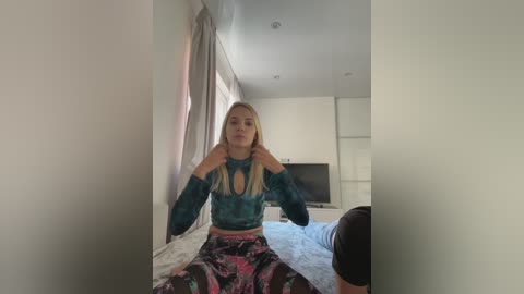 Media: Video of a blonde woman in a teal long-sleeve top and floral-patterned pants, sitting on a bed in a modern, white-walled bedroom with a TV on a dresser in the background.