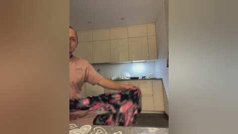 Media: A video of a bald, middle-aged man in a pink shirt and black-and-pink patterned pants, sitting on a kitchen floor, amidst modern white cabinets and a light gray tiled floor.