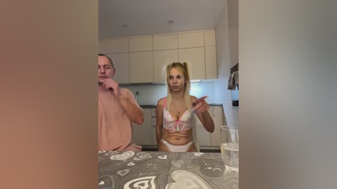 Media: A video of a blonde woman in pink lingerie and a man in casual clothes, standing in a modern kitchen with white cabinets and gray countertops.