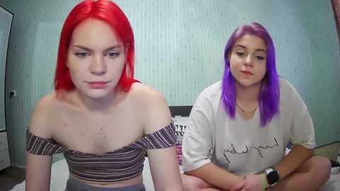 Media: Video of two young women with vibrant red and purple hair, sitting cross-legged on a bed. One wears a striped off-shoulder crop top, the other a white t-shirt with cursive writing. Background shows a light blue, textured wall and a white dresser.