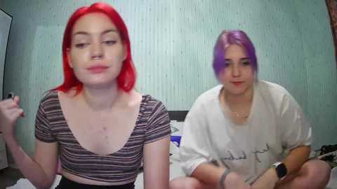 Media: Video of two young women with vivid red and purple hair, one wearing a striped shirt, the other in a white t-shirt, sitting on a bed in a room with light blue textured wallpaper.