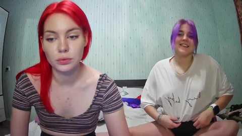 Media: Video of two young women with vibrant red and purple hair, wearing casual clothing, sitting on a bed in a bedroom with light blue wallpaper.