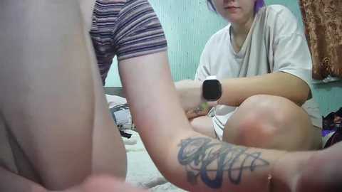 Media: Video of a young person with purple hair, wearing a striped shirt and shorts, using a foot massager on their bare leg with a tattoo visible. Background shows a messy room with teal walls.