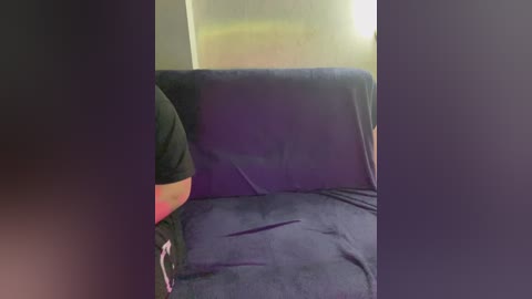 A video of a person sitting on a dark blue sofa, partially obscured by a black curtain. The person's torso and head are visible, wearing a black shirt. The background is a plain, light-colored wall.