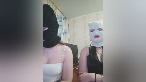 Video of two women, one wearing a black knit mask and a white tank top, the other in a white mask and black top, standing indoors with wallpaper and a TV in the background.