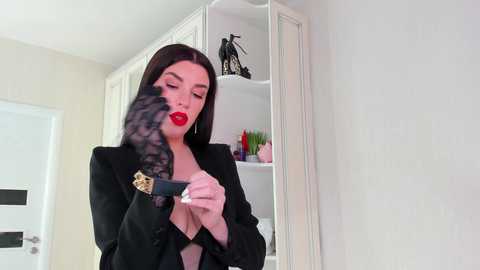 Video of a stylish woman with dark hair, red lipstick, wearing a black blazer, adjusting a black and gold bracelet, standing in a minimalist, white-walled room with shelves holding black sculptures and plants.