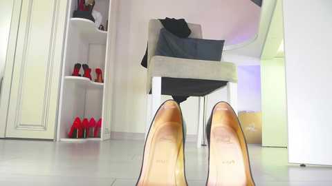A video of a woman's feet in beige high-heeled shoes with red soles, standing in a modern, minimalist room with a grey chair, white shelves, and a mirror.