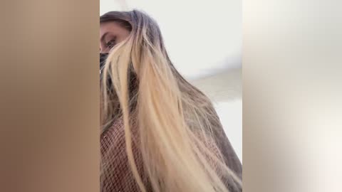 A video of a blonde woman with long, straight hair partially covering her face. She wears a textured, brown sweater. The background is blurred and light-colored.