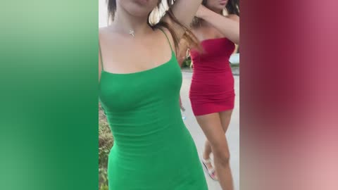 Media: Video of two young women outdoors in the daytime. One wears a tight green dress, the other a red strapless dress. Both have long hair. Background includes blurred greenery and a paved surface.