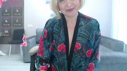 Media: A video of a smiling, blonde-haired woman in a colorful floral robe, standing in a modern, light-colored living room with a grey couch, flowers, and a digital clock on the wall.