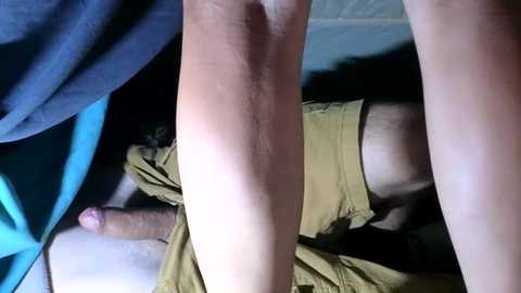Video of a person with light skin and dark hair, lying on their side, showing a bare, erect penis. The person wears a tan shirt and blue fabric. Background includes a blue and white striped pillow.