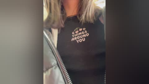 Media: Video of a woman wearing a black T-shirt with the phrase \"COSMIC THINGS AROUND YOU\" in white letters. She has shoulder-length blonde hair and is wearing a white puffy jacket, with blurred background.