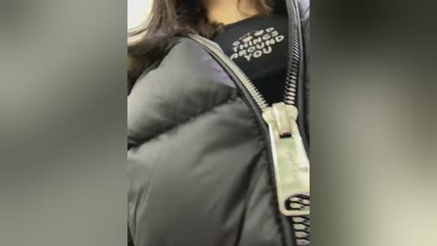 Media: Video of a person wearing a black puffer jacket with a zippered front, partially zipped. The jacket has a white graphic patch with text. The background is blurred, creating a bokeh effect.
