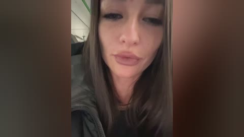 Media: A close-up video of a young woman with light skin, straight brown hair, and full lips puckered in a kiss. She wears a black jacket. The background is blurry, suggesting an indoor setting.