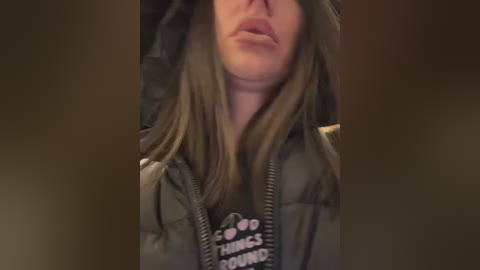 Media: Video of a person with long, straight, light brown hair, wearing a dark green puffer jacket with a graphic design on the chest. Their face is partially obscured, giving a casual, candid vibe.