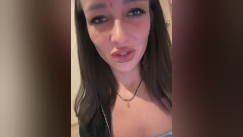 Media: Video of a young woman with long, straight brown hair, fair skin, and blue eyes, wearing a black top and a silver necklace, with blurred background.