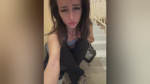 Media: Video of a young woman with long brown hair, wearing a blue tank top and dark pants, sitting on a carpeted floor, looking distressed. Background shows a beige wall and white stairs.