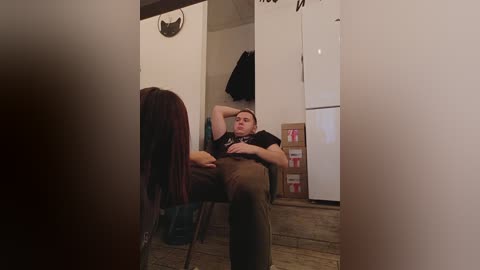 Media: Video of a man sitting on a chair in a dimly lit room with white walls, wooden floor, and a refrigerator. He is wearing a black t-shirt and dark pants.