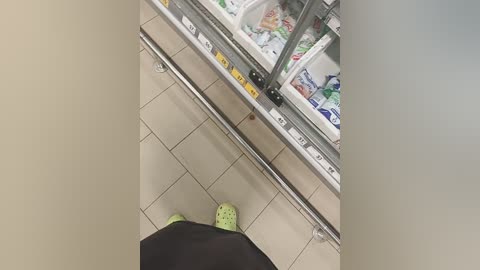 Media: Video of a person in green slippers with black pants, standing in front of a refrigerated grocery display case filled with colorful packaged foods. The floor is tiled in beige.