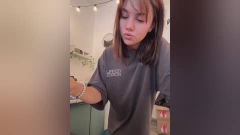 Media: Video of a young woman with medium skin tone and straight brown hair, wearing a gray sweatshirt with a \"United Edition\" logo. She's in a modern kitchen with hanging lights, green cabinets, and a white-tiled backsplash.