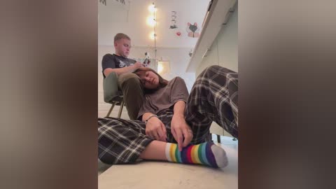 Media: Video of a young man with short hair, wearing a dark t-shirt and plaid pajama pants, sitting on the floor, resting his head on another man's lap.