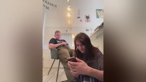 Media: Video of a woman in a salon, sitting, texting, while a man with a beard, wearing a black T-shirt and khaki pants, sits nearby, reading. Background shows white walls, hanging string lights, and framed art.