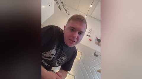 Media: Video of a young, fair-skinned man with short, light brown hair, wearing a black T-shirt featuring a graphic design, standing in a modern, brightly lit room with white walls, displaying artwork.